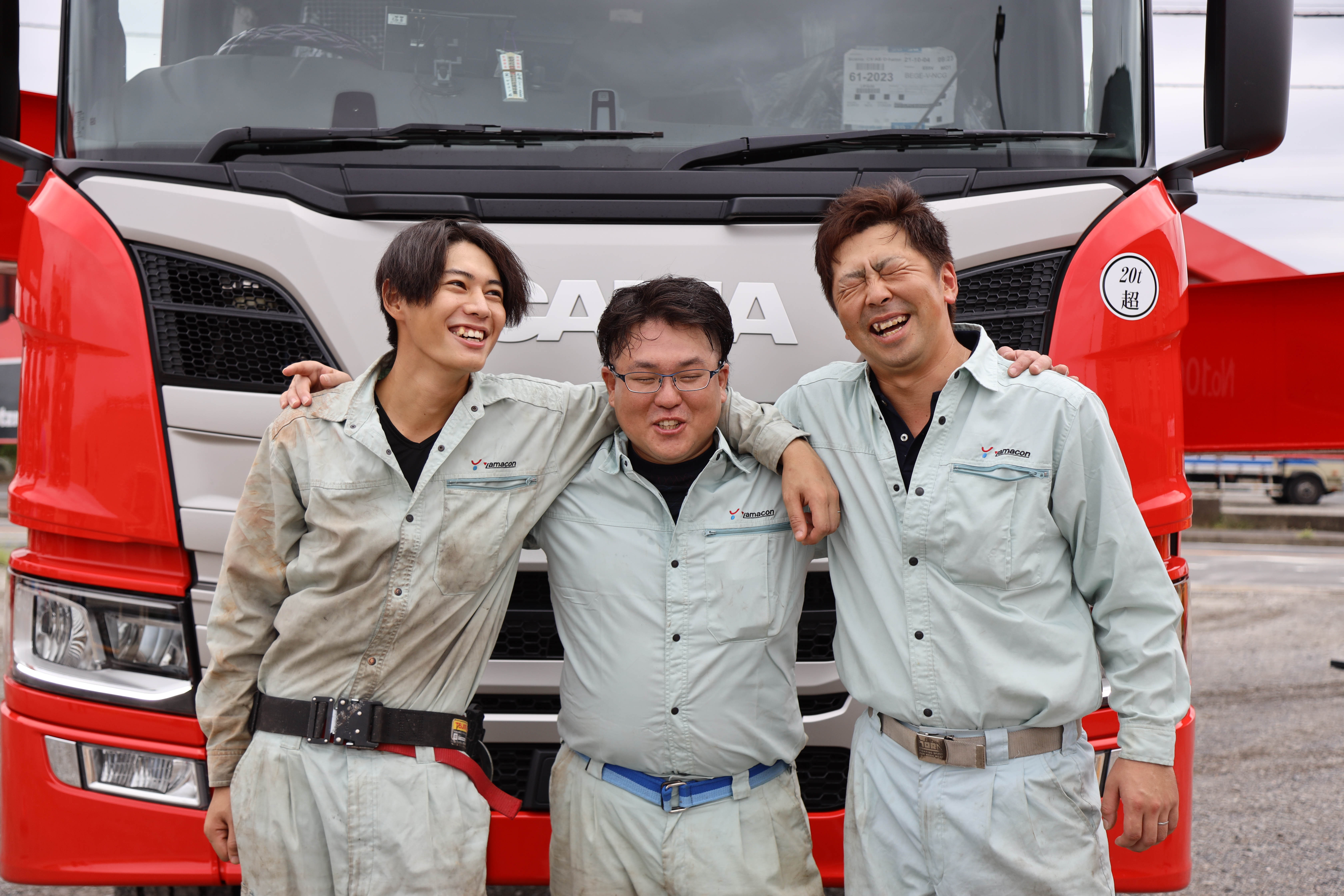 コンクリートポンプ車を背後に作業着の男性3名が肩を組んで並んでいる写真