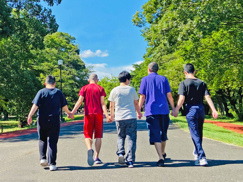 あおいの杜のスタッフと利用者が手を繋いで公園を散歩している姿を後ろから撮った写真