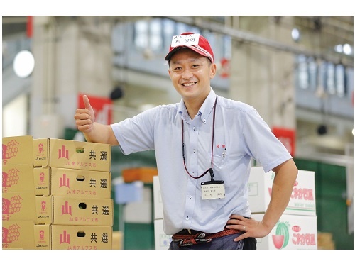積み上げられた果物の段ボール箱の横で、親指を立てて笑顔を見せる赤い帽子の男性。市場のような場所で、爽やかな青いシャツを着て自信に満ちた表情で立っている。
