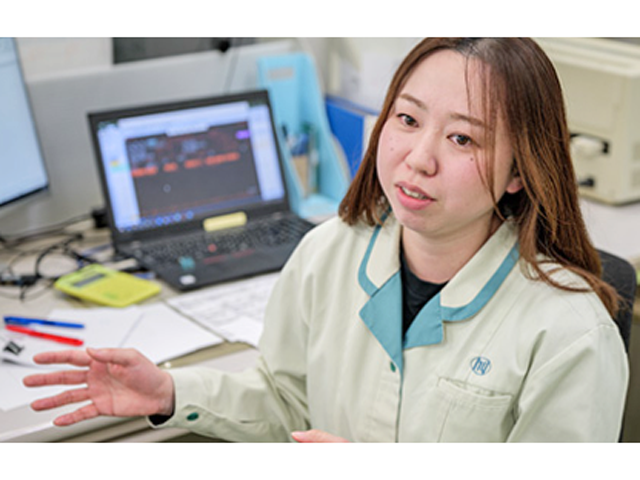 細川洋行　背景にノートパソコンがあるデスクでインタビューを受ける生成りの作業服を着た女性作業員